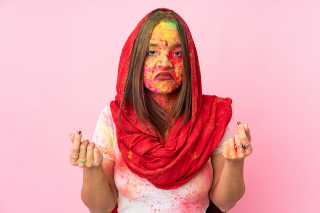 Young Indian woman with colorful holi powders on her face isolated on pink wall making money gesture but is ruined