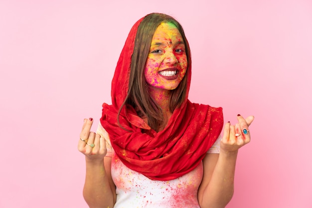 Young Indian woman with colorful holi powders on her face isolated on pink wall making money gesture