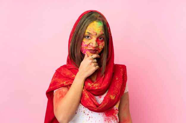 Young Indian woman with colorful holi powders on her face on pink wall thinking