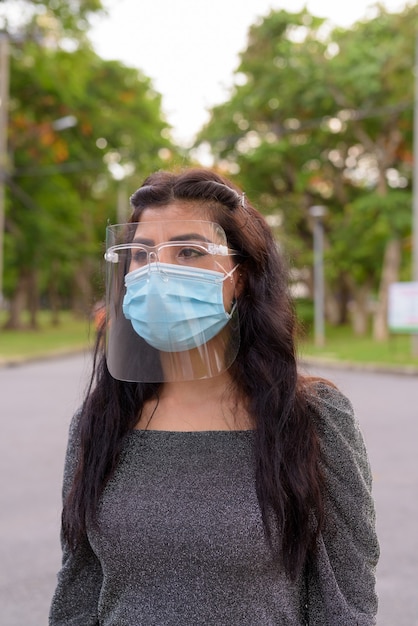 屋外の公園で考えているマスクと顔のシールドを持つ若いインド人女性