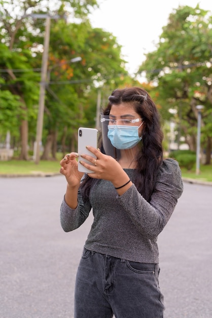 Giovane donna indiana con maschera e visiera utilizzando il telefono presso il parco all'aperto