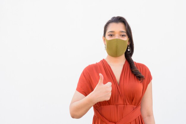 Young Indian woman with mask giving thumbs up against white background