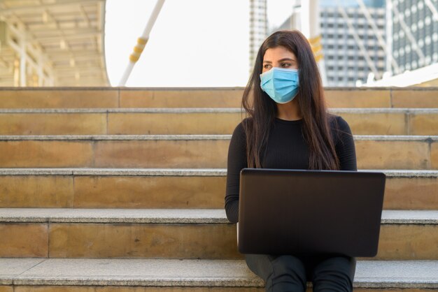 Young Indian woman with mask thinking while using laptop and sitting by the stairs in city