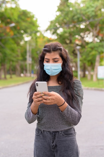 Молодая индийская женщина с маской, используя телефон в парке на открытом воздухе