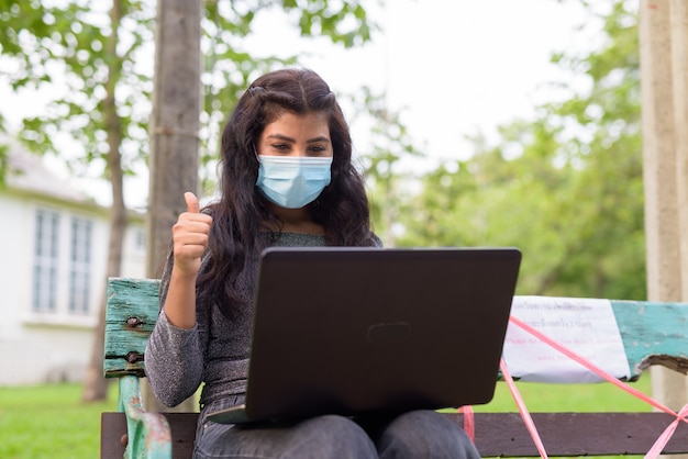 Young Indian woman with mask video calling and giving thumbs up while sitting with distance on park bench