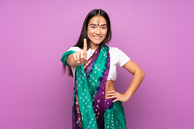 Молодая индийская женщина с сари над изолированной стеной показывая и поднимая палец