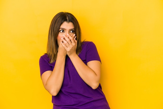 Foto giovane donna indiana su giallo premuroso guardando uno spazio di copia che copre la bocca con la mano.