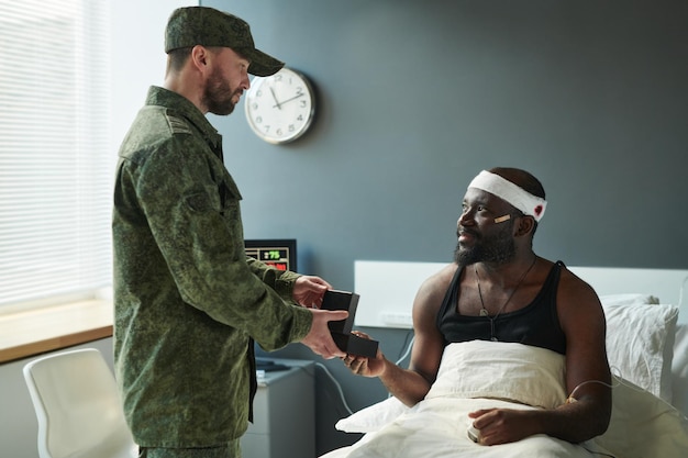 Young injured soldier with bandaged head sitting on bed in front of his friend