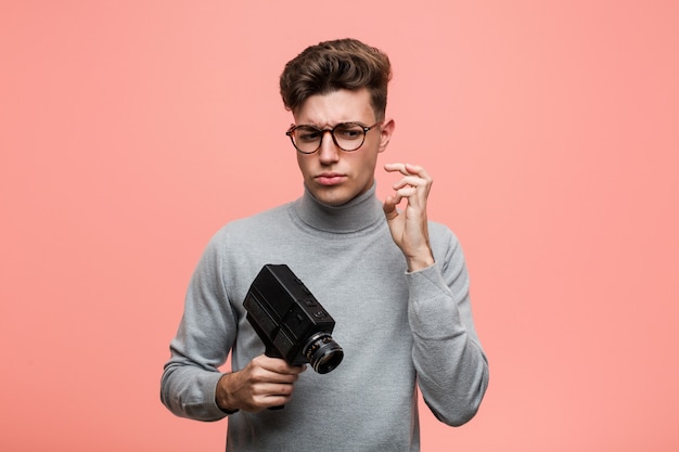 Young intellectual man holding a film camera crossing fingers for having luck