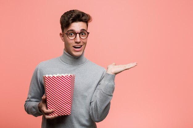 Young intellectual man holding a popcorn bucket cheerful and confident showing ok gesture.