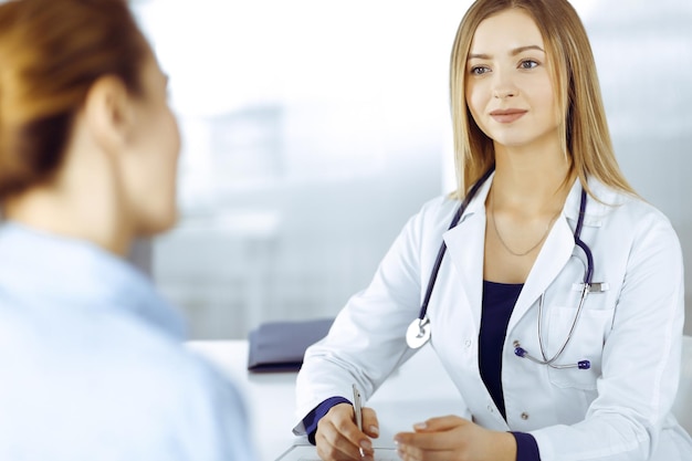 Young intelligent woman-doctor is listening to her patient's allergy's symptoms, while they are sitting together in the cabinet in a clinic. Female physician, with a stethoscope, is writing at clipboa