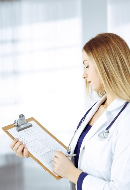 Young intelligent woman-doctor is prescribing some medicine for a patient, using a clipboard, while standing in the cabinet in a clinic. Female physician with a stethoscope at her working place. Perfe