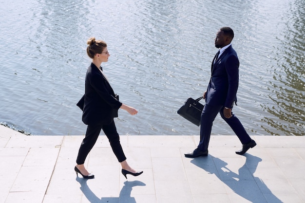 Young intercultural business partners in formalwear moving down\
sidewalk in front of camera along riverside while passing by one\
another