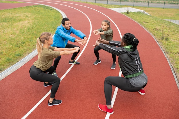 Young intercultural friends in sportswear stretching their arms forwards while doing squats on stadium altogether