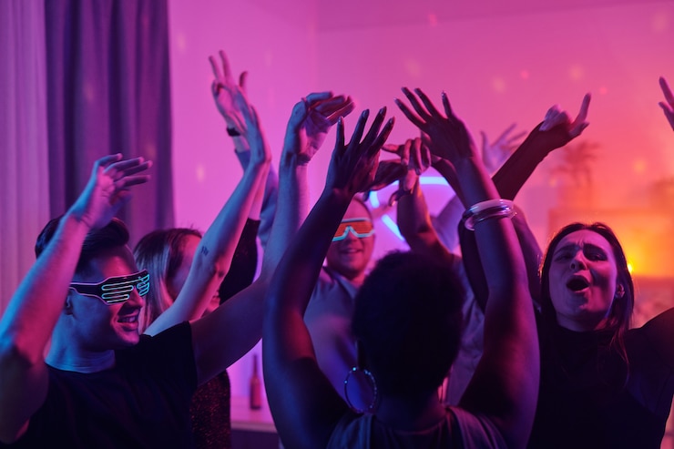 Photo young intercultural glamorous friends raising their arms while dancing together at home party in living-room illuminated with pink lighting