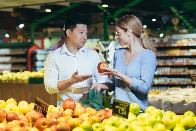 A young international couple a woman and an asian man are choosing fruits in a supermarket deciding