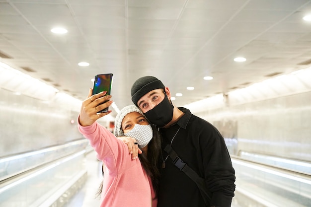 Una giovane coppia interrazziale di amanti che indossano maschere per il viso e cappelli di lana che si fanno un selfie in un corridoio della metropolitana o dell'aeroporto.