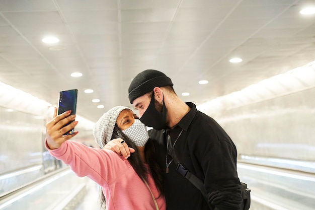 A young interracial couple of lovers wearing face masks and wool hats making a selfie in a subway or airport corridor.