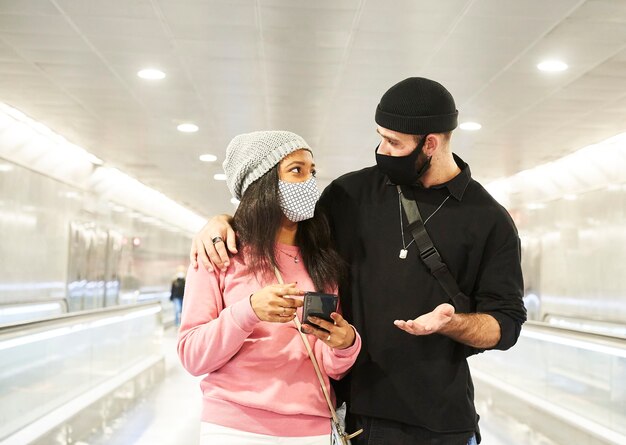 Una giovane coppia interrazziale di innamorati con maschere e cappelli di lana che cammina lungo un corridoio della metropolitana