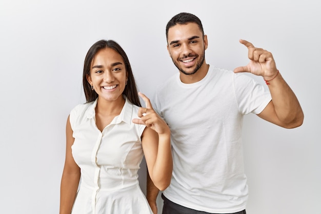 Young interracial couple standing together in love over isolated background smiling and confident gesturing with hand doing small size sign with fingers looking and the camera. measure concept.