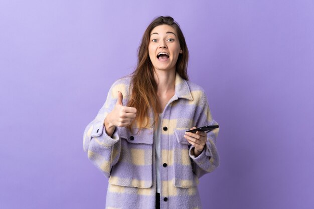 Young Ireland woman isolated on purple wall using mobile phone while doing thumbs up