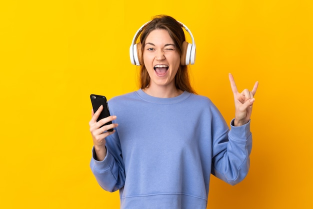 Young Ireland woman isolated on yellow background listening music with a mobile making rock gesture