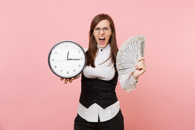 Young irritated business woman in glasses scream hold bundle lots of dollars cash money and alarm clock isolated on pink background. Lady boss. Achievement career wealth. Copy space for advertisement.