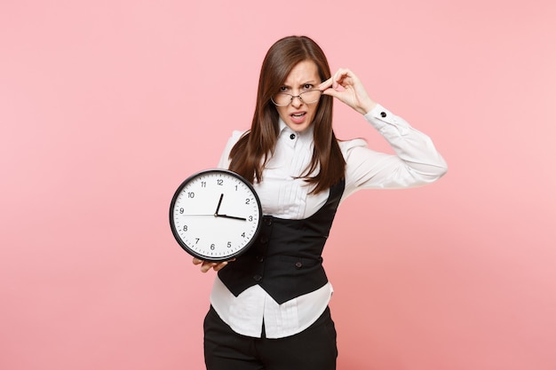 Young irritated dissatisfied business woman in black suit, glasses holding alarm clock isolated on pastel pink background. Lady boss. Achievement career wealth concept. Copy space for advertisement.