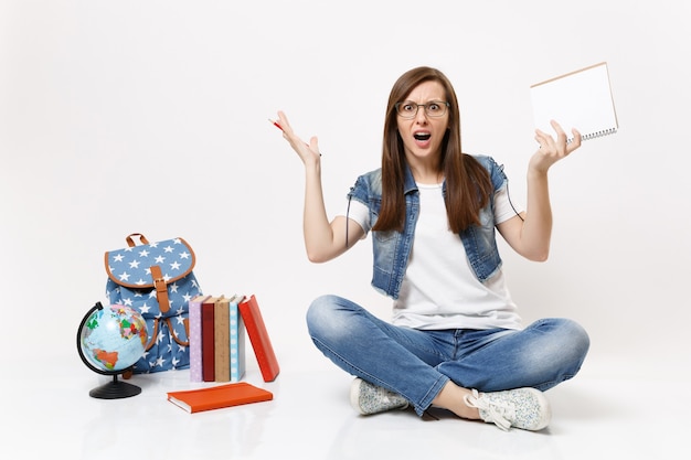 Giovane studentessa irritata con gli occhiali che allarga le mani tenendo una matita, un taccuino seduto vicino al globo, uno zaino, libri scolastici isolati