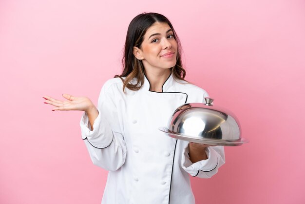 Young Italian chef woman holding tray with lid isolated on pink background having doubts while raising hands