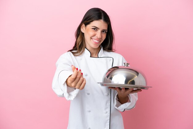 Молодой итальянский шеф-повар женщина держит поднос с крышкой, изолированной на розовом фоне, делая денежный жест