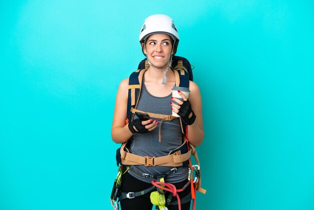Young Italian rock-climber woman isolated on blue background holding coffee to take away and a mobile while thinking something