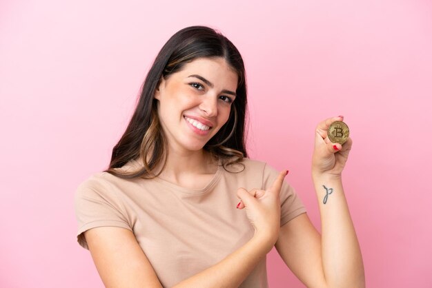 Young Italian woman holding a Bitcoin isolated on pink background pointing back