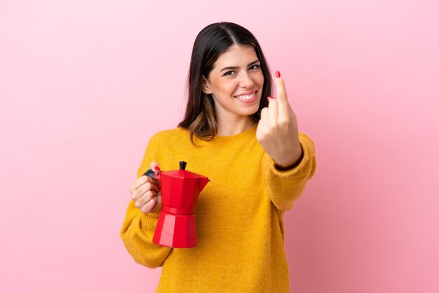 Young Italian woman holding a coffee maker isolated on pink background doing coming gesture