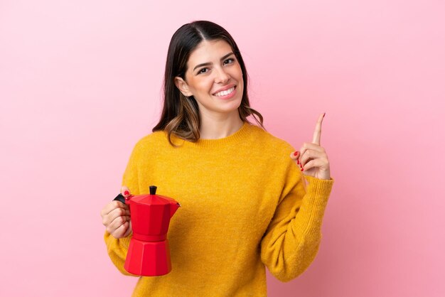 Young Italian woman holding a coffee maker isolated on pink background showing and lifting a finger in sign of the best