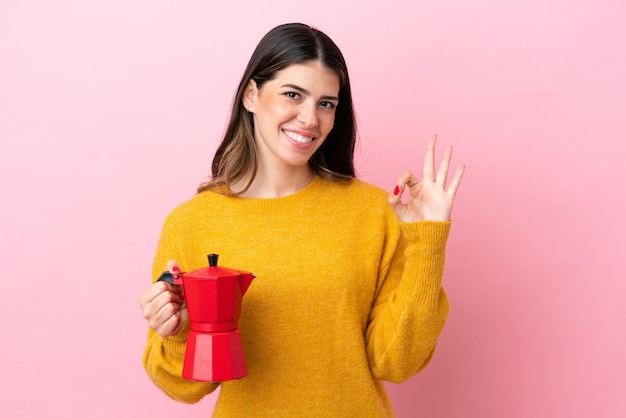 Young Italian woman holding a coffee maker isolated on pink background showing ok sign with fingers