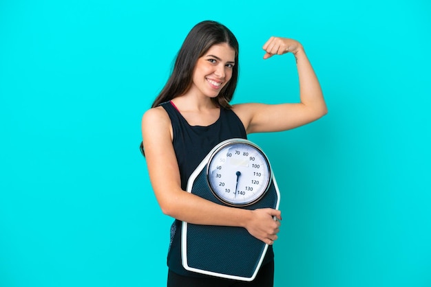 Young Italian woman isolated on blue background holding a weighing machine and doing strong gesture