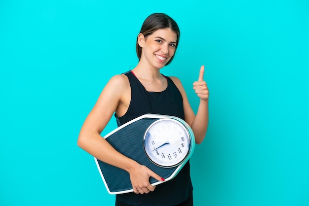 Young Italian woman isolated on blue background holding weighing machine with thumb up