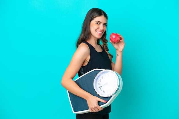 Young Italian woman isolated on blue background with weighing machine and with an apple