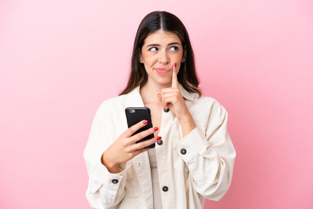 Young Italian woman isolated on pink background using mobile phone and thinking