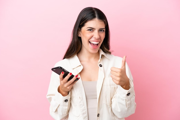 Young Italian woman isolated on pink background using mobile phone while doing thumbs up