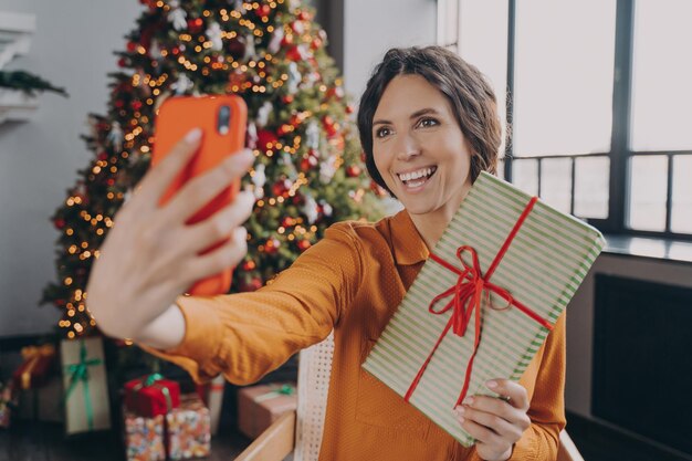 若いイタリア人女性のオンラインブロガーがクリスマスツリーの背景にクリスマスプレゼントで自分撮りを作る