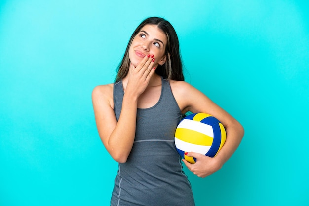 Young Italian woman playing volleyball isolated on blue background looking up while smiling