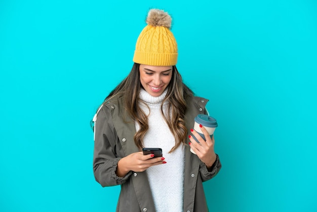 Young italian woman wearing winter jacket and hat isolated on
blue background holding coffee to take away and a mobile