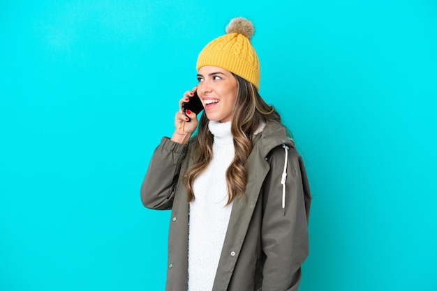 Young Italian woman wearing winter jacket and hat isolated on blue background keeping a conversation with the mobile phone