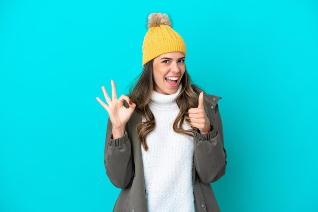 冬のジャケットと帽子を身に着けている若いイタリア人女性は、OKのサインと親指を立てるジェスチャーを示す青い背景で隔離