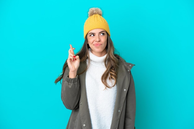 Young Italian woman wearing winter jacket and hat isolated on blue background with fingers crossing and wishing the best