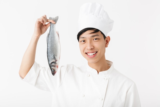 Giovane capo giapponese in uniforme da cuoco bianca e cappello che sorride mentre tiene in mano pesce fresco crudo isolato su un muro bianco