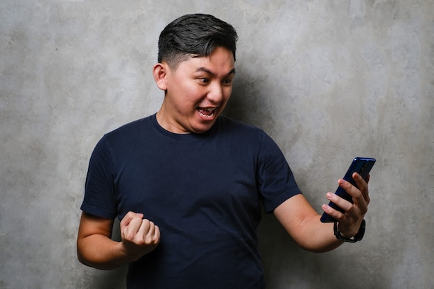 Young japanese man using smartphone standing over concrete wall background screaming proud and celebrating victory and success very excited, cheering emotion