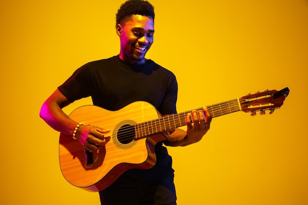 Young and joyful african-american musician playing guitar and singing on gradient orange-yellow studio background in neon light. Concept of music, hobby, festival. Colorful portrait of modern artist.